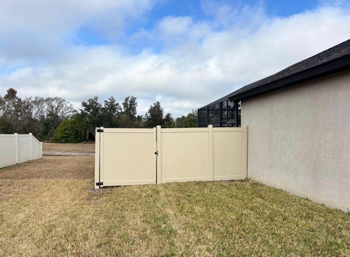 Cream-colored vinyl gate and fence panel in a residential backyard with grass, house, and trees
