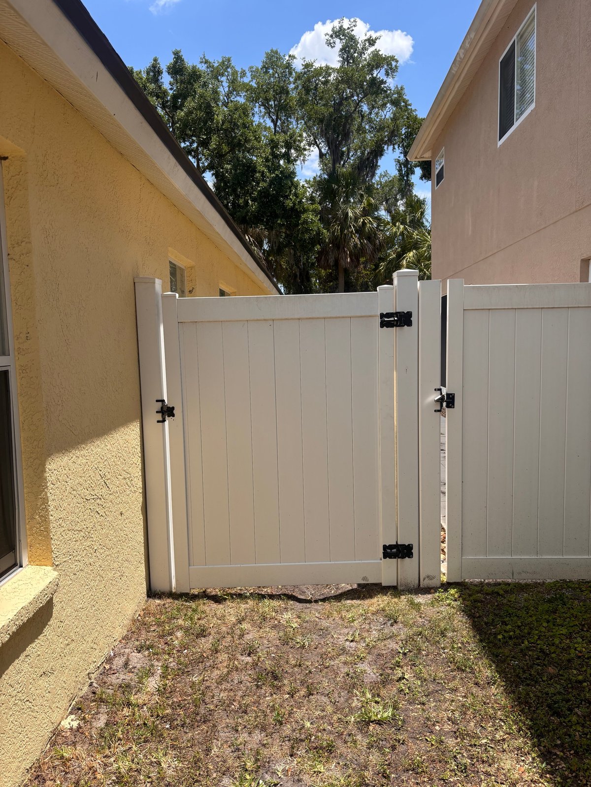 White vinyl gate between yellow and beige house walls with tree canopy visible above against blue sky