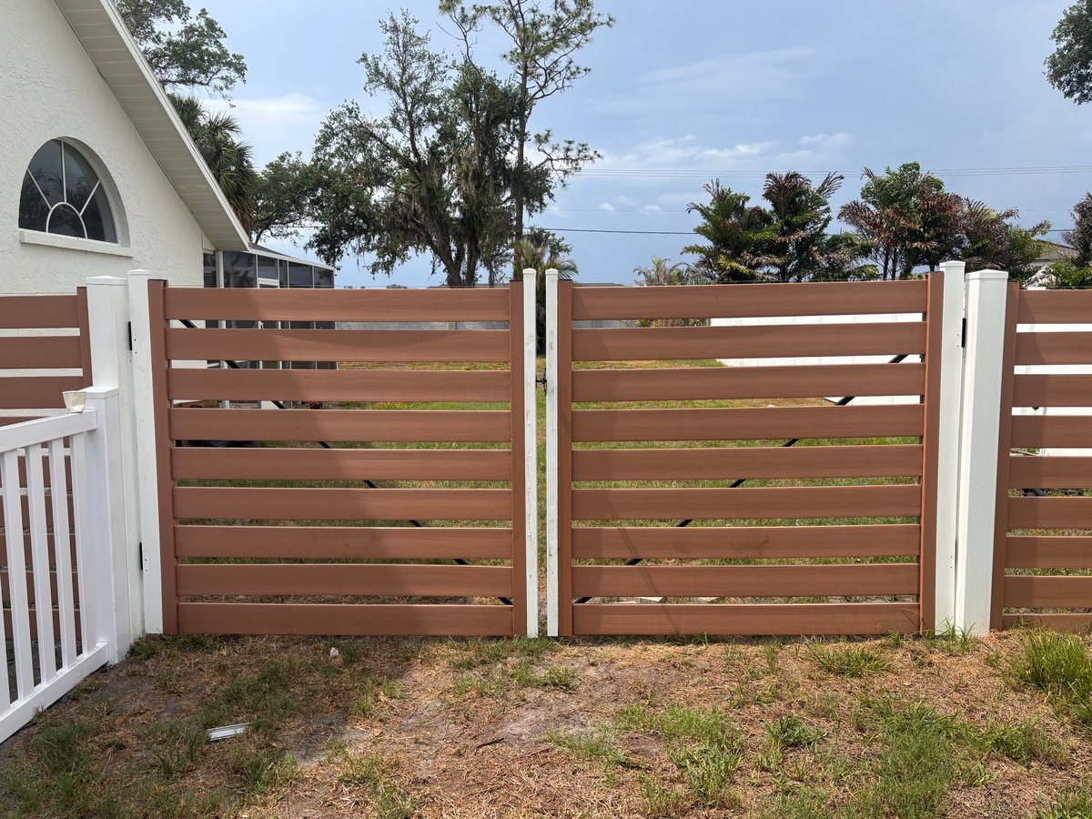 Horizontal wooden fence gate with white posts attached to a house, surrounded by trees and driveway
